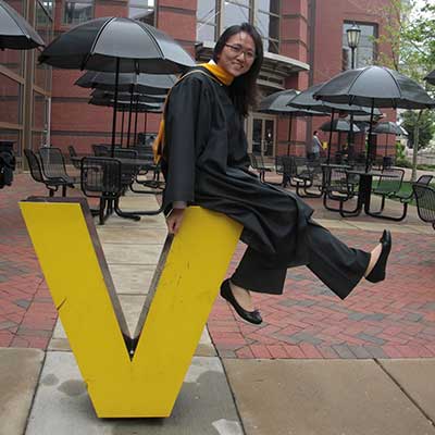 a student dressed in graduation regalia sitting atop a letter v statue on the v.c.u. campus