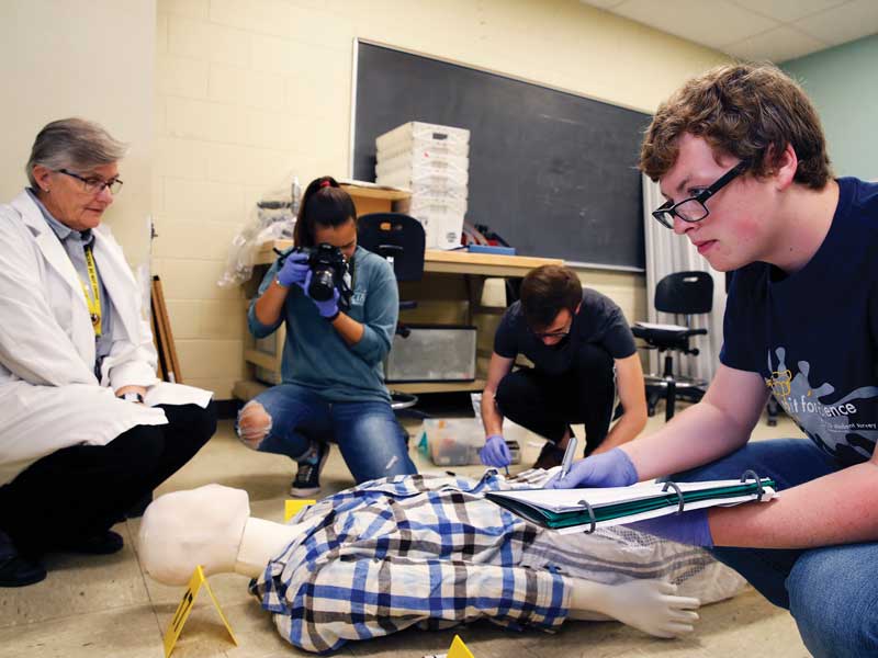 a professor supervises forensic science students conducting an analysis of a fake crime scene
