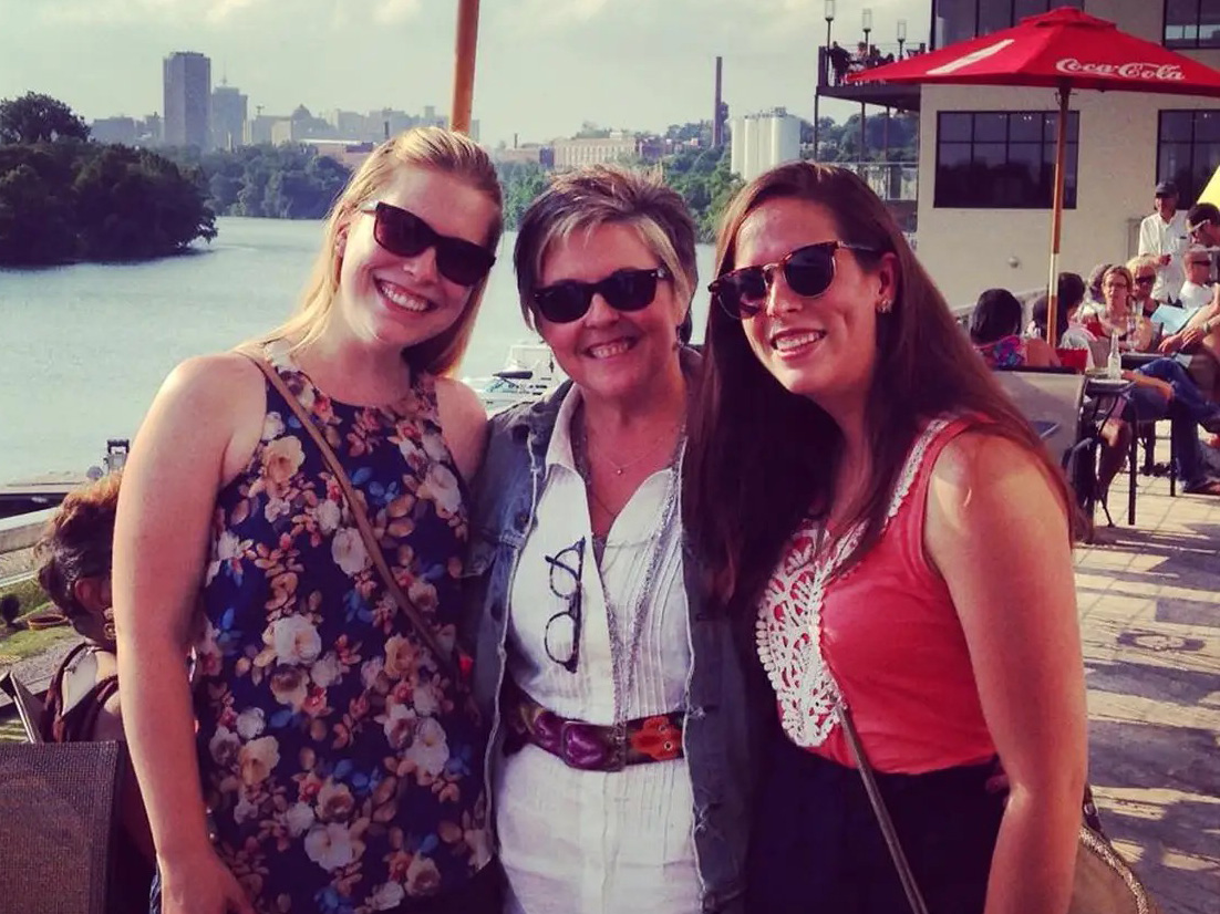 Three women in sunglasses stand at an outdoor restaurant with the James River and Richmond city skyline in the background