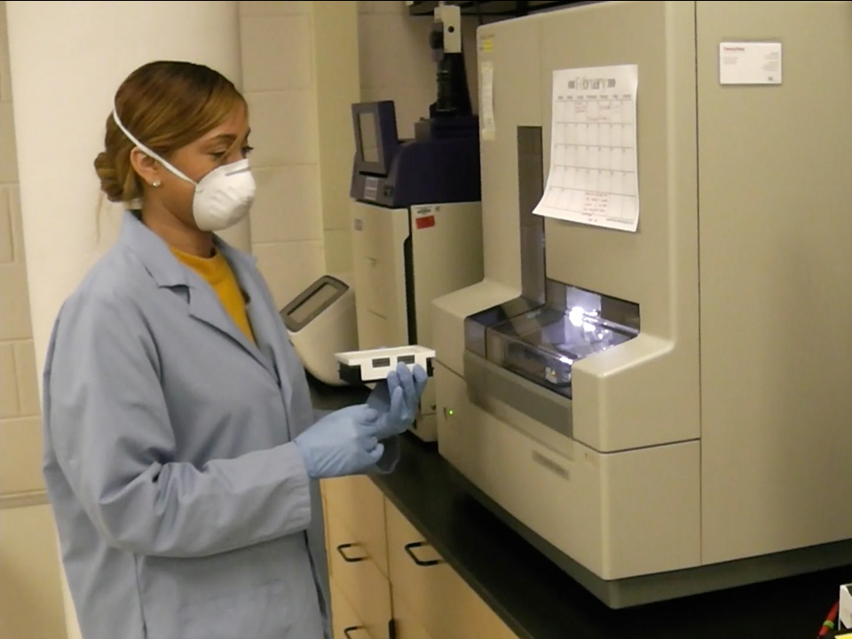 Kelly Knight, wearing a lab coat and face mask, stands in front of equipment in a lab